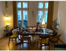 Ein Esszimmer mit einem gedeckten runden Holztisch, umgeben von Holzstühlen mit gelben Polstern. Im Hintergrund sind große Fenster mit gelben Vorhängen, eine Stehleuchte und Zimmerpflanzen sichtbar.