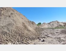 Große Erdhaufen auf einer offenen Fläche mit klarem, blauem Himmel. Im Hintergrund sind weitere Erdhügel und einige grüne Büsche zu sehen.