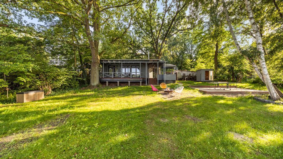 Große, grasbewachsene Gartenfläche mit hohen Laubbäumen und einem modernen, eingeschossigen Holzhaus mit überdachter Veranda und großen Glasfronten. Rechts befinden sich ein befestigter Bereich mit Tischtennisplatte sowie ein kleines Nebengebäude am Rand des Grundstücks.