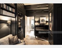 Wohnraum mit dunkler Holzvertäfelung, einer eingebauten Bücherwand und einem schwarzen Klavier. Im Hintergrund sind ein heller Flur und eine teilweise sichtbare Küche zu erkennen, der Boden besteht aus Fischgrätparkett.