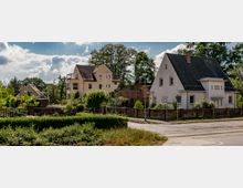 Wohnsiedlung mit Einfamilienhäusern und Gärten in einer ländlichen Umgebung. Die Häuser haben weiße und beige Fassaden, geneigte Dächer und sind von Bäumen und Hecken umgeben.