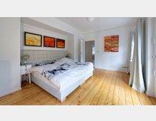 Schlafzimmer mit hellem Holzboden, einem weißen Doppelbett mit gepolstertem Kopfteil und moderner Bettwäsche. An den weißen Wänden hängen abstrakte Kunstwerke in warmen Rottönen, und durch ein großes Fenster fällt Tageslicht herein, das von weißen Vorhängen umrahmt wird.