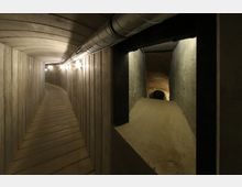 Ein schmaler unterirdischer Tunnel mit Betonwänden und gewölbter Decke, beleuchtet durch eine Reihe von an der linken Wand befestigten Lampen. Rechts führt ein schmaler Durchgang in einen kleineren, sandigen Bereich.