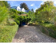Einfamilienhaus mit blauer Ziegeldachdeckung, dessen Fassade von dichter grüner Kletterpflanzenvegetation bedeckt ist, und gepflasterter Zufahrt, die von gepflegten Hecken und Bäumen umgeben ist.