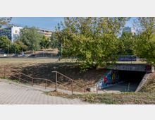 Eine Unterführung mit Graffiti an den Betonwänden, die in eine von Bäumen umgebene Grünfläche eingebettet ist. Im Hintergrund sind eine mehrspurige Straße mit vorbeifahrenden Autos sowie mehrstöckige Wohngebäude zu sehen.