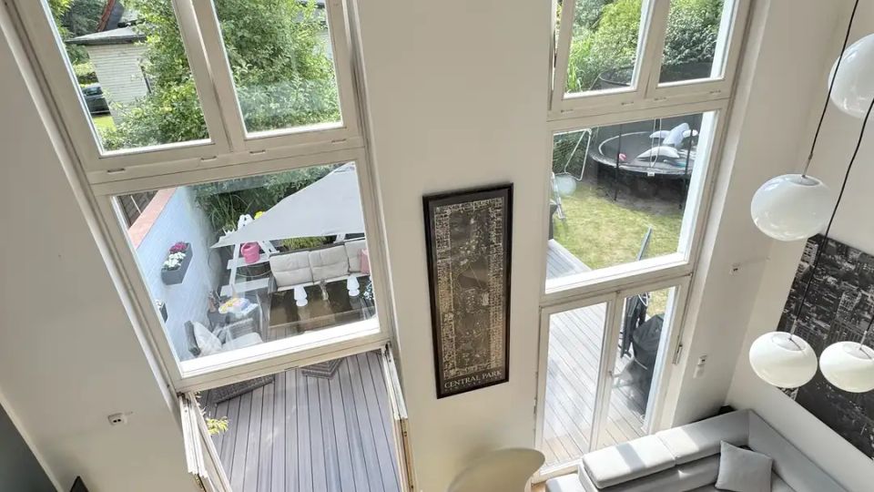 Heller, zweigeschossiger Wohnraum mit hohen Decken, bodentiefen Fenstern und einer Glastür, die auf eine Holzterrasse und in den Garten führt. Im Raum stehen ein graues Ecksofa, ein Couchtisch aus Holz, ein TV-Board und eine hängende Kugelleuchte.