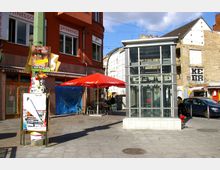 Städtischer Platz mit einer gläsernen Aufzugsanlage im Vordergrund, einem roten Gebäude mit Schildern und einem Café mit roten Sonnenschirmen im Hintergrund. Rechts sind alte Gebäude mit verputzten und unverputzten Fassaden zu sehen.