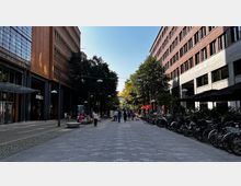 Eine moderne Fußgängerzone mit gepflastertem Weg, flankiert von Ziegel- und Glasgebäuden. Links sind Geschäfte mit Glasfassaden, rechts Fahrradständer und Tische mit Sonnenschirmen, während im Hintergrund Bäume und Passanten erkennbar sind.