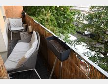 Schmaler Balkon mit dunklem Bodenbelag und einer Bambus-Sichtschutzwand, ausgestattet mit grauen Sitzmöbeln und einem schwarzen Blumenkasten. Im Hintergrund sind grüne Bäume sowie parkende Autos entlang einer Straße sichtbar.