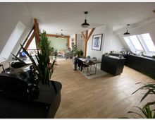 Großer, heller Wohnraum in einer Dachgeschosswohnung mit Holzboden, sichtbaren Holzbalken und schrägen Fenstern. Im Vordergrund ein Klavier und Zimmerpflanzen, im Hintergrund eine Küche mit schwarzen Schränken, ein Esstisch und ein Wohnzimmerbereich mit Sofa und Bücherregal.