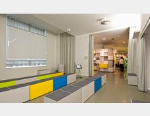 Innenraum eines modernen Museums oder Veranstaltungsraums mit modularen Sitzbänken in Weiß, Gelb, Blau und Grün. Im Hintergrund befinden sich eine Trennwand mit bunten, geometrischen Elementen und einige Personen, die durch den Raum gehen.