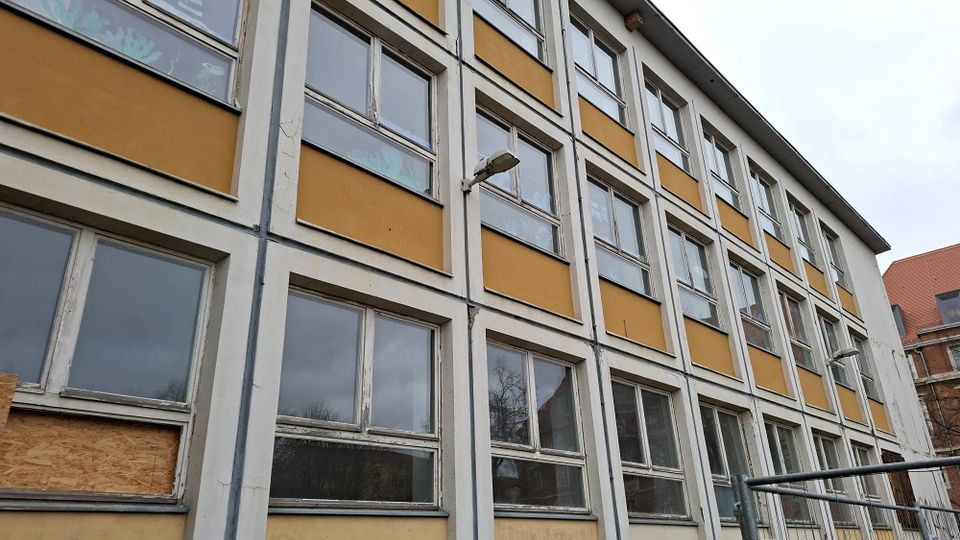 Schräg aufgenommene Fassade eines mehrstöckigen Gebäudes mit gelb-weißen Wandfeldern und vielen rechteckigen Fenstern in gleichmäßigen Rasterreihen. An der Außenwand sind eine Lampe und teils beschädigte bzw. verschlossene Fensterbereiche zu sehen; im Hintergrund ragen Dächer und ein bewölkter Himmel auf.