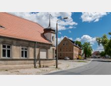 Straßenszene in einer Kleinstadt mit historischen Gebäuden, darunter ein Haus mit rotem Ziegeldach und einem kleinen Turm sowie ein beige verputztes Gebäude im Hintergrund. Die Straße ist asphaltiert und von Kopfsteinpflaster gesäumt; Bäume und blauer Himmel mit Wolken bilden die Umgebung.