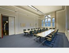 Großer Besprechungsraum mit langen, rechteckigen Tischen und gepolsterten Stühlen in einer klassischen Architektur. Der Raum hat hohe Decken, große Fenster mit Sprossen und helle Wände mit Zierleisten, während durch eine offene Tür ein Korridor sichtbar ist.