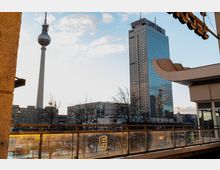 Blick auf den Berliner Fernsehturm und das Hotelgebäude Park Inn mit gläserner Fassade, umgeben von kleineren Gebäuden und kahlen Bäumen im Vordergrund. Eine gläserne Balkonbrüstung mit dem Schriftzug „Balcony“ befindet sich im unteren Bereich des Bildes.