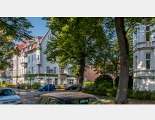 Straßenszene in einer Wohngegend mit mehrstöckigen Gebäuden im Stil der Gründerzeit, weißen Fassaden, grünen Details und roten Dächern. Im Vordergrund stehen Autos geparkt, während Fußgänger auf einem von Bäumen gesäumten Gehweg unterwegs sind.