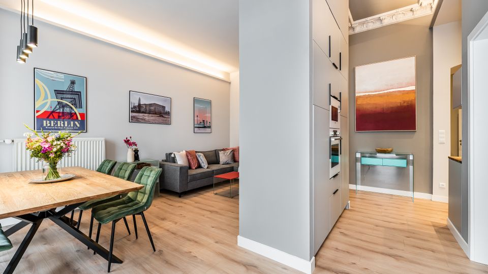 Offener Wohn‑ und Essbereich in einer modernen Wohnung mit Holzboden, Esstisch mit grünen Stühlen, grauem Sofa und gerahmten Wandbildern. Rechts führt ein Flur mit hohen Einbauschränken und integrierten Geräten zu einem Konsolentisch unter einem großen abstrakten Gemälde.