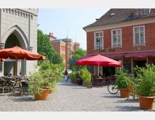 Straßenszene mit Kopfsteinpflaster, einem Café mit roten Sonnenschirmen und Sitzmöglichkeiten im Freien, umgeben von grünen Pflanzen in Töpfen. Im Hintergrund sind ein Gebäude mit gotischem Bogen und eine historische Backsteinfassade sichtbar.