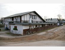 Ein einstöckiges Gebäude im Fachwerkstil mit weiß gestrichenen Wänden und braunen Holzelementen, ergänzt durch ein schräges Dach. Im Vordergrund sind ein Fahrrad, ein Fenster mit Blumenkasten und ein unbefestigter, sandiger Untergrund sichtbar.