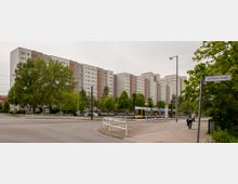 Breite Wohngebäude im Stil des Plattenbaus mit mehreren Stockwerken, ergänzt durch Grünflächen mit Bäumen im Vordergrund. Eine Straßenbahn hält an einer Haltestelle, und ein Straßenschild mit der Aufschrift "Jan-Petersen-Straße" ist sichtbar.