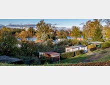Kleingartenanlage mit mehreren kleinen Lauben und Parzellen, umgeben von Bäumen und Sträuchern in herbstlichen Farben, im Hintergrund ein See und einzelne größere Gebäude am Horizont.