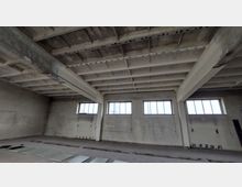 Ein leerer, industrieller Innenraum mit Betonwänden und einer Betonrippendecke. Der Raum hat mehrere rechteckige Fenster mit einfachem Glas und Rahmen an der oberen Wand, die mattes Tageslicht hereinlassen, und es liegen Platten auf dem Boden verteilt.