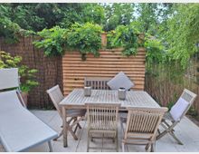 Ein kleiner Gartenbereich mit einer Terrasse aus Steinplatten, ausgestattet mit einem Holztisch und passenden Stühlen. Im Hintergrund eine Sichtschutzwand aus Holz und Bambus mit Kletterpflanzen sowie umliegenden Sträuchern und Bäumen.