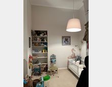 Ein Kinderzimmer mit weißer Wand, einem kleinen weißen Gitterbett, einer Spielzeugaufbewahrung und Regalen, die Bücher und Stofftiere beherbergen. An der Wand befinden sich kindgerechte Dekorationen wie eine Giraffenfigur und ein gerahmtes Bild, auf dem ein Teddybär abgebildet ist.