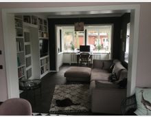 Wohnzimmer mit Ecksofa und Teppich, angrenzend an einen offenen Arbeitsbereich mit Schreibtisch und Stuhl vor einem großen Fenster. An der linken Wand befinden sich Regale mit Büchern und ein eingebauter Fernseher.