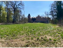 Blick über eine große, grasbewachsene Wiese auf ein historisches Schloss aus rotem Backstein mit Türmen und Zinnen. Das Gebäude liegt am Waldrand und wird von hohen, teils kahlen Bäumen eingerahmt, darüber ein klarer blauer Himmel.