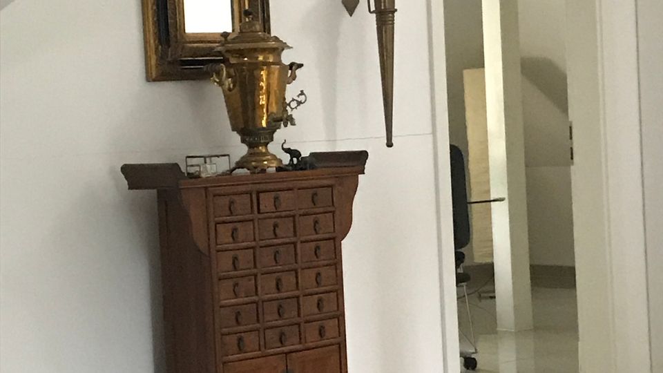 Innenansicht eines hellen Flurs mit glänzendem Fliesenboden und schräger Decke. An der Wand steht ein hohes Holzschränkchen mit vielen kleinen Schubladen, darüber ein gerahmter Spiegel und eine Wandlampe; rechts führt eine offene Tür in einen weiteren Raum.
