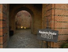 Eingangsbereich eines historischen Gebäudes mit gemauerten Backsteinwänden und einem gepflasterten Boden. Im Vordergrund steht ein hölzernes Schild mit der Aufschrift „Keramik-Studio Dorothea Nerlich“.