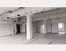 Ein leerer Raum in einem renovierungsbedürftigen Gebäude mit weißen Fliesen an den Wänden, mehreren offenen Durchgängen und losen Kabeln, die von der Decke hängen. Der Boden ist teilweise aufgerissen, und die Beleuchtung besteht aus freiliegenden Neonröhren.