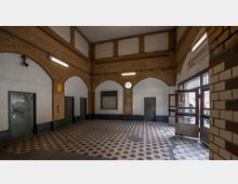 Innenraum eines historischen Bahnhofseingangs mit Backsteinmauern und weißen Wandflächen, bogenförmigen Tür- und Fensteröffnungen sowie einem rot-weiß gemusterten Boden. Eine analoge Uhr hängt zentral an der Wand, und rechts führen Glastüren nach draußen.