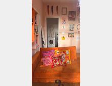 Wohnzimmer mit orangefarbener Couch, die mit farbenfrohen Kissen mit Mustern und Totenkopfmotiven dekoriert ist. Im Hintergrund ist ein weißer Bereich mit einer Waschmaschine und einer Schaufensterpuppe zu sehen, umgeben von pastellrosa Wänden mit Bildern, Masken und Rahmen als Wanddekoration.