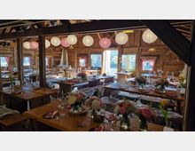 Rustikale Scheunen-Gaststube mit sichtbaren Holzbalken und Holzvertäfelung, ausgestattet mit langen Holztischen und Bänken, die mit Fellen belegt sind. An der Decke hängen runde Papierlaternen, und die gedeckten Tische sind mit Gläsern, Besteck sowie Blumenvasen dekoriert; Tageslicht fällt durch mehrere Fenster mit rosa Vorhängen.