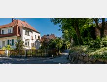 Straßenszene mit einem zweistöckigen Wohnhaus mit rotem Satteldach und grünen Fensterläden auf der linken Seite, einem Holzzaun im Vordergrund und einem gepflasterten Gehweg, der an einer mit Bäumen und einer alten Mauer bewachsenen Grünfläche entlangführt. Im Hintergrund sind weitere Ziegeldächer und ein Straßenschild erkennbar.