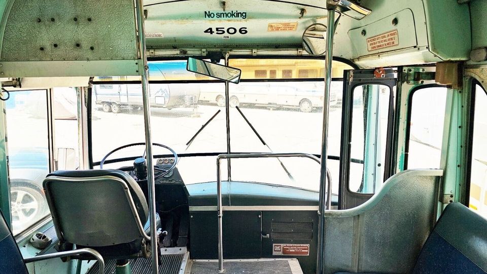 Innenraum eines alten Linienbusses mit Blick auf den Fahrersitz und das Armaturenbrett. Die Sitzpolster und Wände sind in Grün- und Blautönen gehalten, und vorn sind Schilder zu sehen, darunter eines mit der Aufschrift „No smoking“.