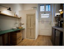 Kleine Küche mit Holzboden, einer hölzernen Arbeitsplatte, offenen Regalen und Küchenutensilien an der Wand. Rechts sind eine weiße Tür, ein Fenster und eine Kaffeemaschine zu sehen.