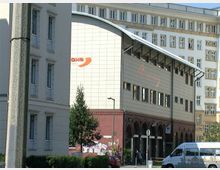 Straßenansicht eines modernen, mehrstöckigen Gebäudes mit hellen Fassaden, großflächigen Fenstern und einem geschwungenen Dach. Im Vordergrund stehen ältere Wohnhäuser, ein Baum und ein geparkter Minivan.