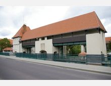 Brücke mit integrierten Gebäudeteilen und orangefarbenem Ziegeldach, umgeben von Geländern und umgeben von Bäumen. Die Konstruktion zeigt Elemente aus Stahl und Putz sowie Fensteröffnungen im oberen Bereich.