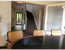 Esszimmer mit einem runden Tisch und Stühlen mit Geflechtlehnen im Vordergrund. Im Hintergrund ein Flur mit Treppe und Glastür, die nach draußen führt.