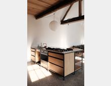 Moderne Küche mit einer freistehenden Kücheninsel aus Holz und Metall, ausgestattet mit einem Gasherd, einem Backofen und Schubladen. Im Raum sind sichtbare Holzbalken an der Decke, ein großer Papierlampenschirm und eine helle, minimalistische Einrichtung zu sehen.