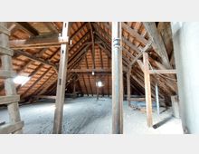 Dachboden mit sichtbarem Holzbalkenwerk und schrägen Dachziegeln an der Innenseite. Der Raum ist staubig, mit Holzboden und kleinen Dachfenstern, die Licht einlassen.