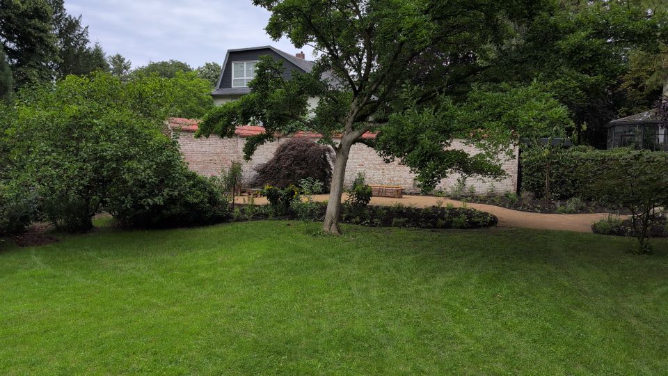 Gartenanlage mit großer Rasenfläche, einem zentral stehenden Baum und umlaufenden Beeten mit Sträuchern. Im Hintergrund verläuft eine hohe Ziegelmauer mit roter Abdeckung, dahinter ragt ein Wohnhausdach hervor; rechts ist ein kleiner Gewächshausbau zu sehen.