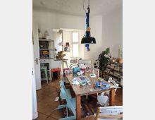 A dining room or multi-purpose space with a wooden table cluttered with dishes, books, and papers, surrounded by mismatched chairs. The room features white walls, wooden flooring, a large hanging black light fixture, tall windows, shelves filled with magazines, and a large potted plant.