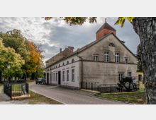 Historisches Gebäude mit hellem Putz und Satteldach, ergänzt durch einen kleinen Turm mit spitzem Dach, neben einem gepflasterten Weg und einem Wasserrad im Vordergrund. Im Hintergrund sind Bäume mit Herbstlaub und ein wolkiger Himmel sichtbar.