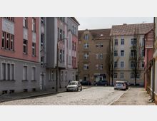 Straße in einem Wohnviertel mit mehrstöckigen Altbauten in pastellfarbenen Fassaden, die mit dekorativen Stuckelementen verziert sind. Die gepflasterte Straße ist von parkenden Autos und spärlich belaubten Bäumen gesäumt.