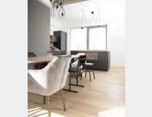 Ein moderner Essbereich mit hellem Holzboden, einem rechteckigen Holztisch und verschiedenen Stühlen, darunter ein grauer Samt-Sessel. Im Hintergrund befindet sich eine minimalistische Küche mit dunklen Fronten, großen Fenstern und einer Hängelampen-Installation.