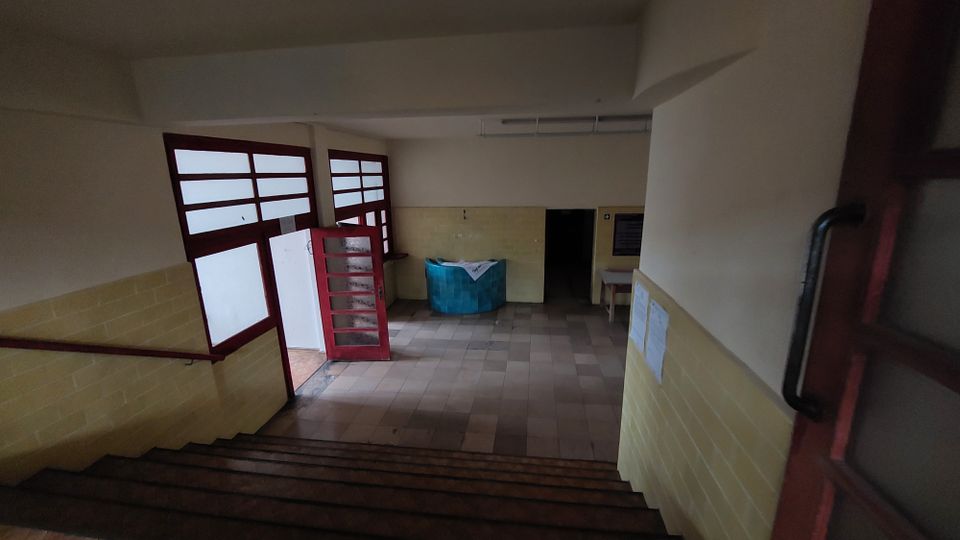 Treppenhaus in einem schlichten Gebäude mit hellgelb gefliesten Wänden und einem Boden aus beigen Fliesen. Unten führen rote Türen aus Holz mit Glaselementen in einen weiteren Raum, in dem ein türkisfarbener runder Empfangstresen steht.